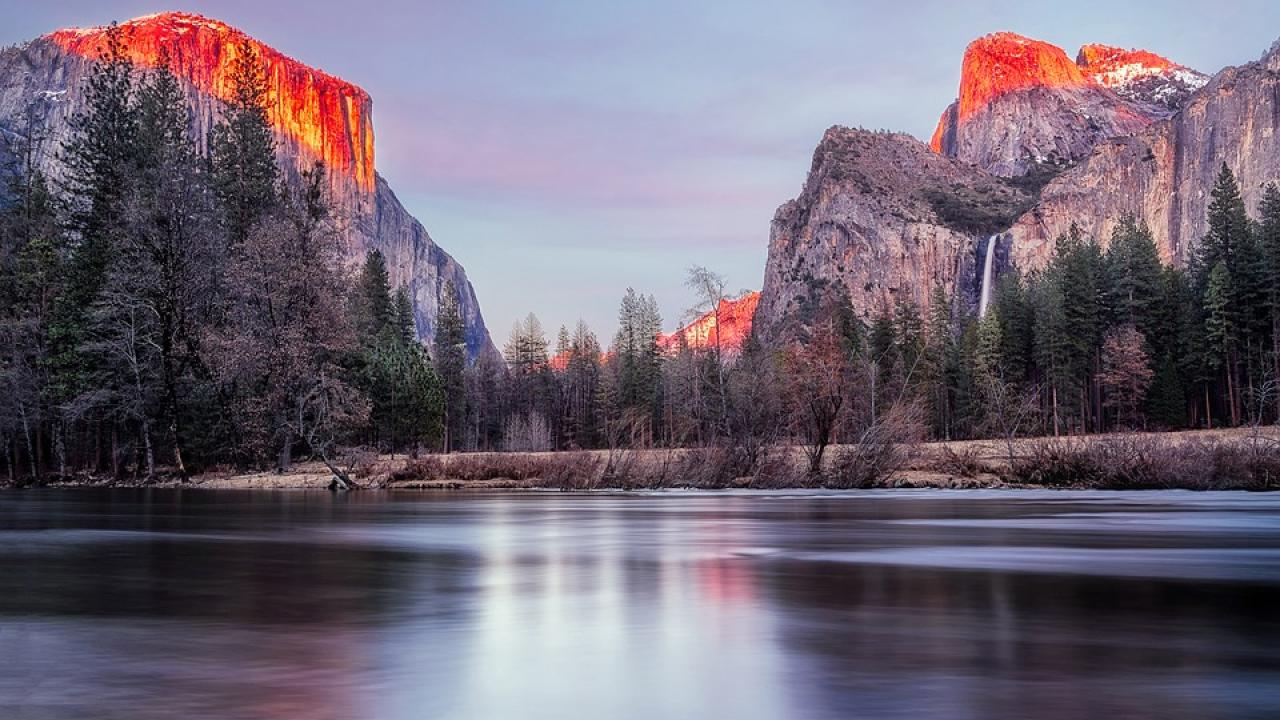 Image: a lake at dusk flanked by mountains and trees.